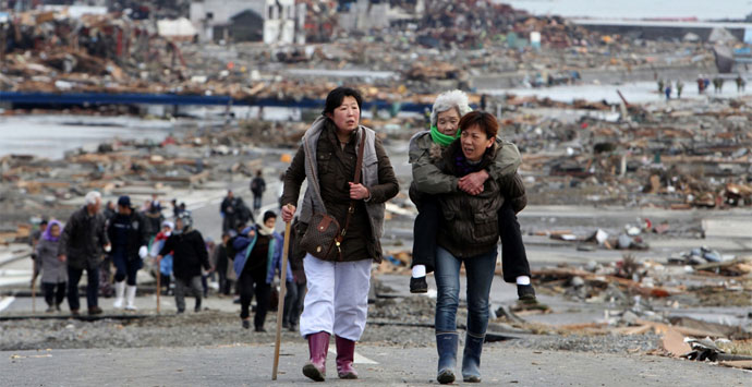 A woman carries an elderly woman on her back after the 2011 tsunami in Japan. Minami-Sanriku-cho, a town located in Miyagi Prefecture is unrecognizable. Credit: The Kahoku Shimpo.